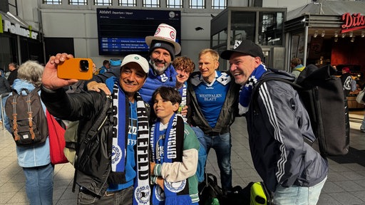 Eine Gruppe von Arminia Fans jeden Alters steht in einem Bahnhof
