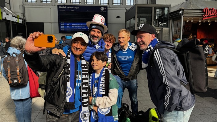 Eine Gruppe von Arminia Fans jeden Alters steht in einem Bahnhof