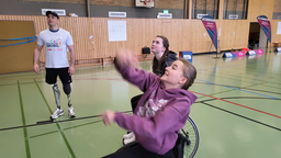 Zwei Mädchen im Rollstuhl und ein Junge mit Beinprothese spielen zusammen Basketball in einer Sporthalle.