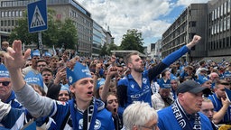 Viele hunderte Arminia-Fans, zwei reißen die Arme in die Luft. 