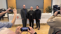 Drei Männer in Anzügen werden von der Presse fotografiert