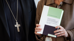 Eine Frau hält rechts im Bild ein Buch in der Hand, links neben ihr steht ein Mann in einem schwarzen Gewand und goldener Kreuzkette.