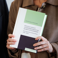 Eine Frau hält rechts im Bild ein Buch in der Hand, links neben ihr steht ein Mann in einem schwarzen Gewand und goldener Kreuzkette.