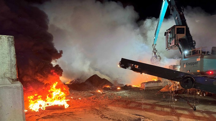 Flammen auf dem Recylinghof. Im Rauch steht ein Bagger.