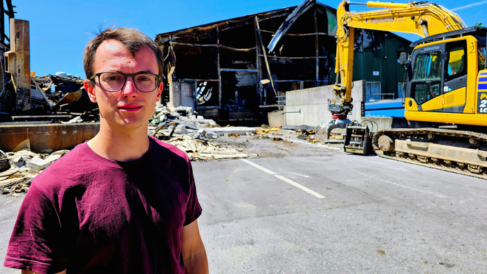 Ein Mann mit Brille steht vor einer abgebrannten Halle.