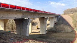 Über ein ausgeschachtetes Tal mit braunem Geröll erstreckt sich der Neubau einer Betonbrücke mit einem seitlichen Windschutz aus rotem Plexiglas.