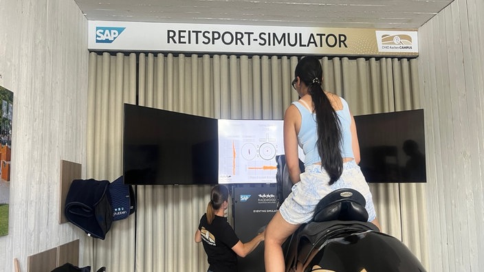 Frau sitzt auf schwarzem Plastik-Pferd vor einem Schild Reitsport-Simulator