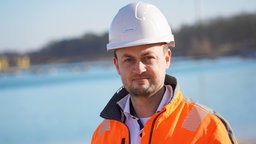 Ein Mann mit Warnjacke und Helm steht vor einem See