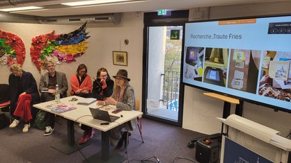 Mehrere Frauen sitzen an einem Tisch und zeigen auf einer Leinwand eine PowerPoint zur Entstehung des Buches.