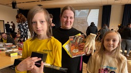 Drei Personen stehen nebeneinander und gucken in die Kamera. Sie halten Dinge wie eine Hand aus Plastik und Comichefte in der Hand