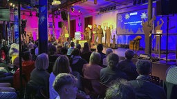 Moderator:innen stehen auf der Bühne
