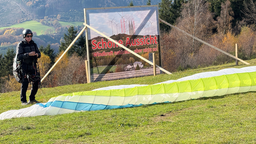 Ein Gleitschirmflieger steht auf einer Wiese. Dahinter ein Plakat auf dem "Schöne Aussicht. Windkraft natürlich aber nicht so!" steht.