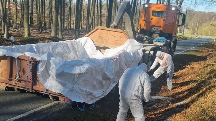 Zwei Mitarbeiter in weißen Schutzanzügen stehen neben einem Nutzfahrzeug am Straßengraben.