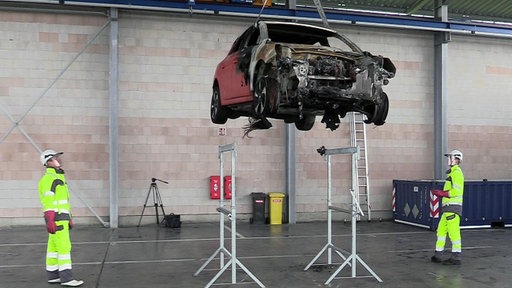 Ein abgeschlepptes E-Auto wird in einer Werkstatt hochgefahren und von zwei Sicherheitsmenschen begutachtet.
