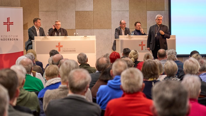 Erzbistum Paderborn. Zwei Männer stehen auf der linken Seite hinter einem Rednerpult. Auf der rechten Seite befindet sich ebenfalls ein Rednerpult mit drei Männern. Vorne sitzt das Publikum.