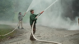Zwei Männer stehen mit Wasserschläuchen vor einem Haus und besprühen dieses mit Wasser. 