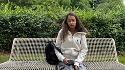 Die Freundin des Opfers sitzt auf einer Bank und schaut in die Kamera. Sie hat ein Handy in der Hand und trägt eine graue Strickjacke.