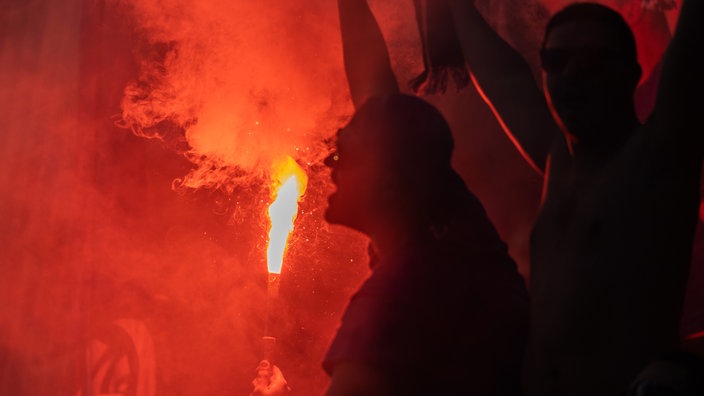 Ein bengalisches Feuer, eine rot leuchtende Fackel mit weißer Rauchentwicklung, beleuchtet von hinten den dunklen Kopf eines Mannes.