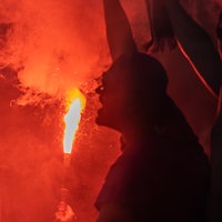 Ein bengalisches Feuer, eine rot leuchtende Fackel mit weißer Rauchentwicklung, beleuchtet von hinten den dunklen Kopf eines Mannes.