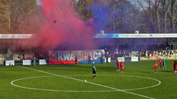 Ein Fußballstadion mit Zuschauern auf der Tribüne und Spielern auf dem Feld. Ein rot-blauer Qualm steigt in die Luft.