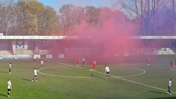 Ein Fußballstadion mit Spielern auf dem Feld. Links tragen die Spieler weiße Trikots, rechts rote. Ein rot-blauer Qualm verdeckt die Zuschauer auf derTribüne. 