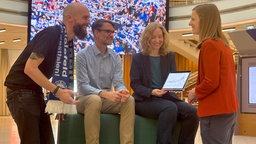 Vier Menschen unterhalten sich. Eine Frau zeigt eine Grafik und ein Mann trägt einen Arminia Bielefeld-Schal. Im Hintergrund sieht man das Spiel auf einem Bildschirm.