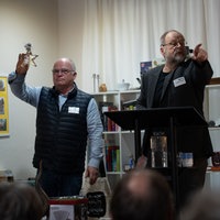 Zwei Männer stehen auf einer Bühne. Einer hält ein Objekt aus Glas hoch. Der andere deutet mit dem Finger in die Menschenmenge vor ihnen.