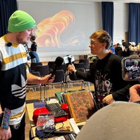 Zwei Personen unterhalten sich über einem Tisch hinweg. Die Linke Person hält ein Mikrofon, die rechte Film sich während des Gesprächs selbst. Eine dritte Person filmt die beiden beim Gespräch mit einem Handy