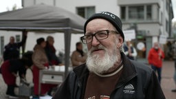 Ein älterer Mann mit weißen Bart lächelt in die Kamera, hinter ihm wird Suppe ausgegeben. 