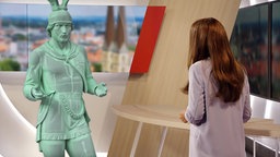 Man sieht einn KI-generiertes lebensgroßes Hermannsfigur im Lokalzeit OWL-Fernsehstudio im Interview mit der Moderatorin.