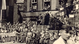 Bild aus dem Jahr 1950 vom Detmolder Marktplatz. Man sieht viele Menschen, die vor dem Rathaus sitzen und auf der Treppe stehen. Sie applaudieren, lachen und freuen sich. Das ist ein Bild der Feier anlässlich des 75. Geburtstags des Hermannsdenkmals