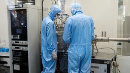Zwei Forscher in blaun schutzanzügen stehen an einem Forschungsgerät