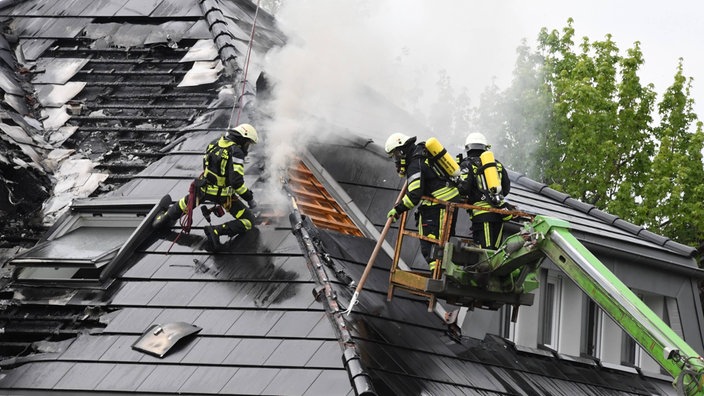 Drei Feuerwehrleute löschen einen rauchenden Dachstuhlbrand an einem beschädigten Wohnhaus.