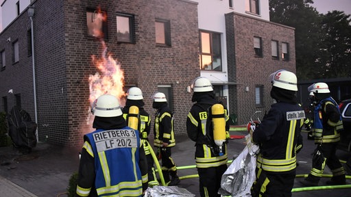 Mehrere Feuerwehrleute stehen vor dem brennenden Haus und schauen auf eine Stichflamme, die gerade aus einem Kellerfenster kommt und an der Fassade empor steigt