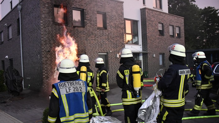 Mehrere Feuerwehrleute stehen vor dem brennenden Haus und schauen auf eine Stichflamme, die gerade aus einem Kellerfenster kommt und an der Fassade empor steigt