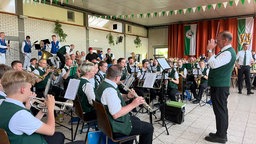 Zahlreiche Musiker in einer mit Bannern geschmückten Halle spielen in weißen Hemden und grünen Westen ihre Instrumente.