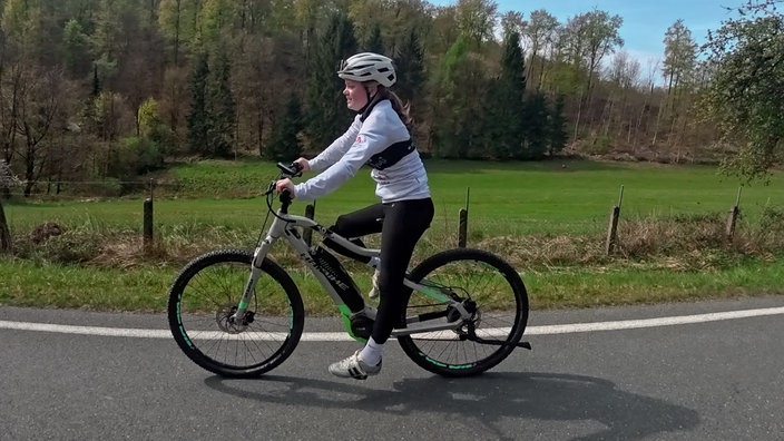 Anni Wein fährt mit dem Fahrrad auf einer Straße vor einer grünen Wiese
