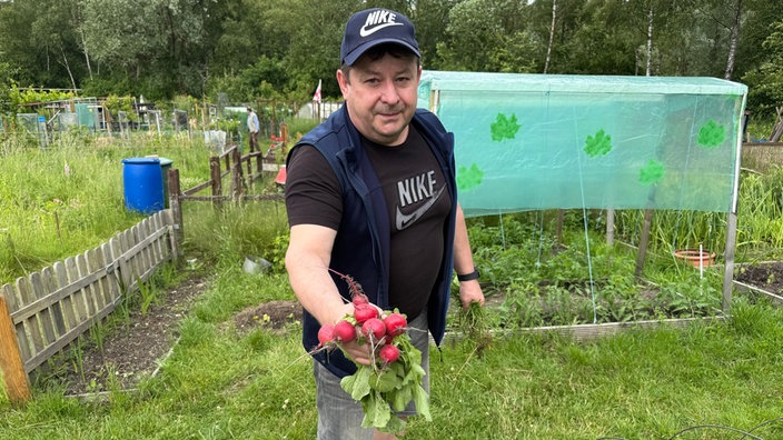 Ein Mann steht in einem Garten und hält einen frisch geerntetes Bund Radieschen in die Kamera.