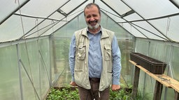 Ein Mann steht mitten in einem Gewächshaus und lacht. Auf dem Boden sind verschiedene Pflanzen in der Erde.