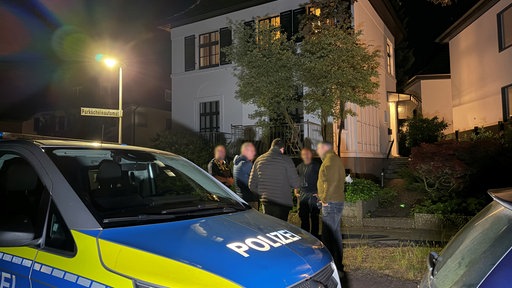 Mehrere Männer stehen nachts vor einem Haus, daneben steht ein Streifenwagen der Polizei.