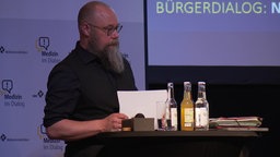 Ein bärtiger Mann, Jörg Schrader, steht auf einem Podium hinter einem kleinen Tisch