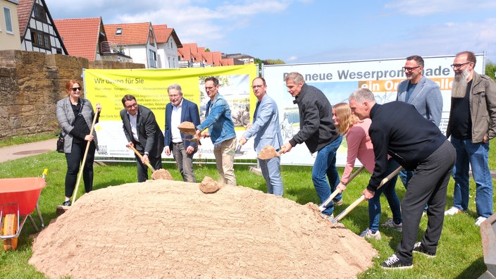Politiker und Verwaltungsmitarbeiter:innen mit Spaten vor einem aufgeschütteten Sandhaufen beim symbolischen Spatenstich