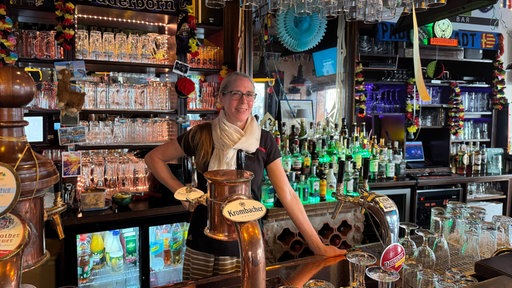 Eine Mitarbeiterin lehnt hinter dem Tresen einer dekorierten Sportsbar und lächelt in die Kamera. Die Bar ist mit vielen Gläsern und Getränken ausgestattet.