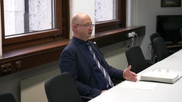 Oberstaatsanwalt sitzt vor einem Ordner mit vielen Blättern und spricht