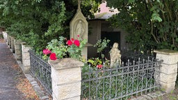 Ein Bildstock mit grünen Pflanzen, roten Rosen und zwei Steinskulpturen am Rande eines Weges