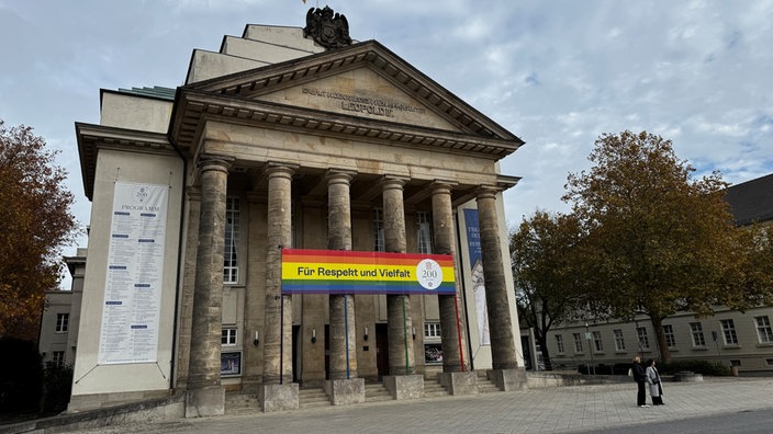 Auf dem Bild sieht man das Landestheater wie es heute aussieht. Mit sechs hohen Säulen vor dem Eingang. An den Säulen hängt ein Banner in Regenbogenfarben mit der Aufschrift "Für Respekt und Vielfalt" anlässlich des 200. Jubiläums.