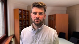 Ein Mann in Hemd steht in einem Raum mit Holz-Bücherregalen und blickt in die Kamera. 