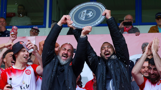 Mehrere Personen jubeln in einem Stadion. Zwei Männer halten eine große silberne Schale mit der Aufschrift "FLVW" hoch.