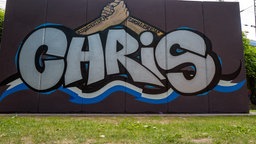 Ein Graffitibild an einer Wand, welches den Namen Chris zeigt. Darüber sind zwei Arme zu sehen. Die Hände halten sich fest.