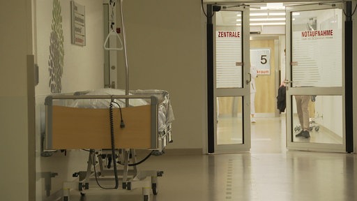 Der Gang zu einer Notaufnahme eines Krankenhauses. Direkt vor dem Eingang zur Notaufnahme steht ein leeres Krankenbett.